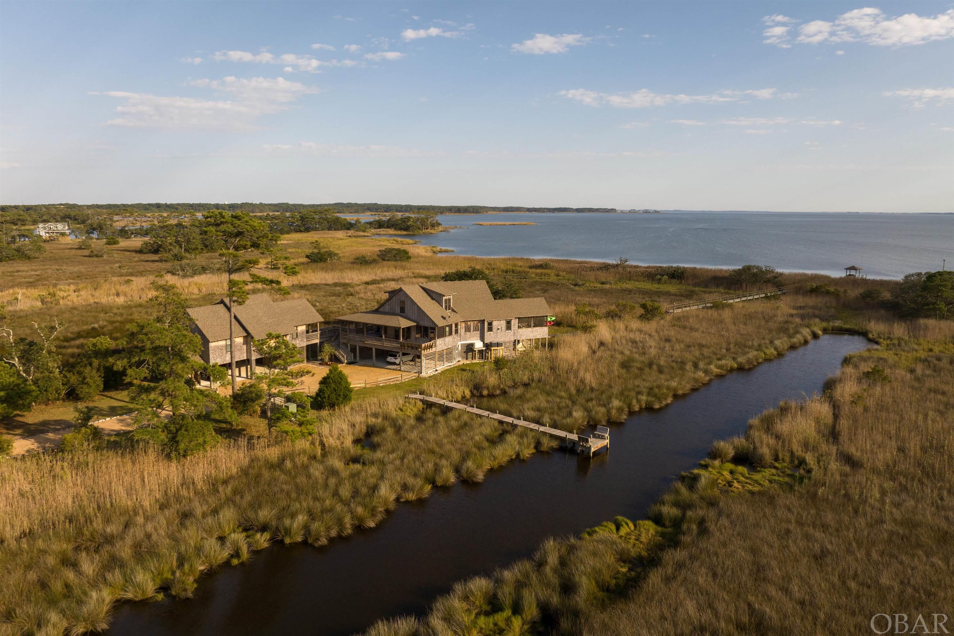 If you've been searching for a private retreat surrounded by nature, look no further than Breezy Point, a captivating 26-acre SOUNDFRONT gated estate just three miles from the Atlantic Coastline in Kill Devil Hills.  This custom-built property, featuring a gorgeous MAIN residence and DETACHED CARRIAGE HOUSE, was designed to meld seamlessly with its natural surroundings, offering a sense of calm, comfort and quietude to all who enter.  Opportunities to connect with nature abound, with wildlife such as deer, rabbits, and otters as well as waterfowl and marine life spotted frequently.  After nightfall, the property is enrobed in starlit skies and moonlit waters.  Both the main residence and carriage house were designed to take full advantage of the coastal scenery.  The main residence features reclaimed and refinished yellow pine flooring throughout and consists of either 3 or 4 bedrooms (depending on your desires for the space) and 2.5 baths, with designer details and luxurious touches throughout the space.  The main level offers a bright and open living area featuring a gas fireplace with custom yellow pine surround, custom window seats with built-in storage, and access to a massive soundfront screened porch.  The gourmet kitchen features new stainless appliances, durable and elegant soapstone countertops, custom cabinetry, and a walk-in pantry.  There is also a fabulous pass-through butler's pantry that transitions to the dining room, which also offers al fresco dining thanks to another adjacent screened porch.  Wake up to dazzling sound views from the primary suite, which is conveniently located on the main level and features built-in window seating with storage, an attached screened porch, and a luxurious private bath with heated marble tile flooring, tiled shower, double sink vanity with marble top, and easy access to both the adjacent walk-in closet and laundry room.  Depending on your needs, upstairs, you will find two to three additional guest rooms along with a shared bath with double sink vanity and custom tiled bath surround, all situated around a spacious loft with marsh and canal views.  The detached carriage house is absolutely gorgeous, with reclaimed and refinished oak flooring, 15-foot ceilings, designer lighting, and transom windows.  This space can be tailored to fit a variety of needs, such as a guest cottage, gameroom and media lounge, home office, or artisan's studio.  The kitchenette can be easily expanded into a full kitchen thanks to a capped gas line in the east wall.  This space also offers expansive storage, luxurious full bath, covered parking and private entry, and its own whole home generator, along with sprawling views of the marshes and sound beyond.  There is no shortage of outdoor living area, with covered porches, screened porches, and sun decks ensconcing the perimeter, along with a massive ground level lounge space perfect for crab picking or a lowcountry boil at sunset with friends.  Meander through the marsh grasses on the private concrete walkway to take in breezes on the soundfront pier and gazebo, or read a book or catnap in the secluded screened pond house.  Launching your kayak, paddleboard or boat is easily accomplished at the private boat dock situated along the canal.  From the custom architectural details, to the breathtaking views, to the many opportunities to connect with nature, this unique, expansive property is a picturesque private paradise.
