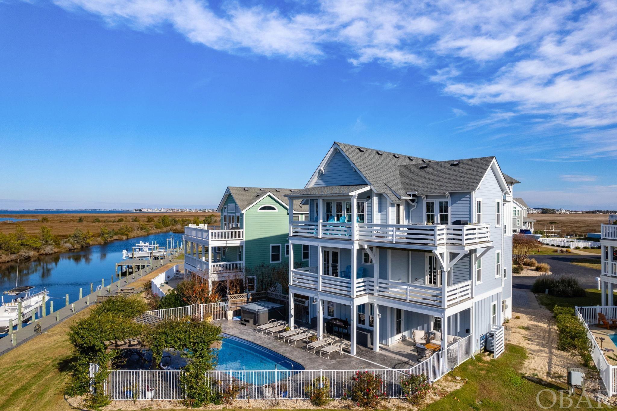 100 Sunset Ct. is the epitome of all things Coastal! The location is certainly coastal, as it’s located in the small waterfront community of the Peninsula in Manteo, NC. There’s 140ft. of deep-water bulkheaded canal available at your back door to dock your boat/boats/jet skis/ pontoon or kayaks! The bulkheaded walkway encircles the community and offers access to the navigable Shallowbag Bay channel connecting the Roanoke Sound and Pamlico Sound through the Oregon Inlet to the Atlantic Ocean.  The home was designed by Michael York of The Coastal Cottage Co.  Sitting atop a high lot, there are terrific views of the canal, the sound and marsh so that morning, noon and sunset time, you’re surrounded by brilliant blues of sky and water or cloud colors indicative of the boating weather for the next day. This home has immediate curb appeal from the driveway pavers to the cottage architecture.   A beautiful custom front door and sidelights allow light into the welcoming foyer providing privacy and light to the mid-level where you’ll find Eddie Bauer engineered hardwood floors, coastal trim and details all round. In the hallway, there’s an elevator, closet and laundry with washer/ dryer and an extra freezer for your “catch of the day”.This functional space is neatly disguised by sliding barn door for easy access. There are 3 bedrooms on this level, all with balcony/deck access. There’s a primary bedroom on the north west with a generous closet and large, private bath. The double vanity with the glass vessel sinks is flanked on one end with a private water closet on one side and a large walk-in tiled shower with transom window, built in seating and shower heads on the other. A linen closet completes the picture. There are two other bedrooms on this level which are connected by a Jack and Jill bath with 2 separate/private vanities with vessel sinks, bench seating as well as separate closets for each. The linen closet holds everything you need for enjoying the large walk-in tiled shower with a transom window. The two bedrooms on the north side have canal views and the bedroom on the south looks out across the community to the sound beyond . Take the elevator up to an open concept top floor with vaulted ceilings and hand crafted beams. The spacious kitchen has room for more than one cook or separate prep areas with more cabinet spaces than you could ask for. The 48”gas cook top with pot filler, double electric oven, dishwasher, wine cooler are all connected by leathered granite countertop and has ample seating all round for those who like to watch the cook and get first dibs on the feast. The light fixtures in this area are made from Handcrafted California wine barrel rings. Gather around the gas fireplace with a live edged mantle central to the seating area and a generous separate dining area adjacent to the one of the best butler’s pantries you’ve seen in a while. There’s room for a slim extra fridge and it’s all accessed by a sliding barn door. Step out to an open west facing deck where you can see the sound and Jockey’s Ridge to the east or sunsets to the west. There’s also another bedroom on this level which is currently used as an office/sleeping area. The full bath with tile shower makes it a very comfortable, accessible spot. Take the elevator/stairs down to the ground level with luxury plank floors which is great when you’re coming into the house from a swim or grilling on the spacious pool deck. The large salt water pool is really the icing on the cake! It’s surrounded by a generous pool deck large enough for several lounge chairs and the beautiful jasmine covered trellis. Access to the canal is easy via 3 fence gates so you easily jump on the boat, come back for a swim and soak in the 7-person hot tub or grab a refreshing beverage for a game of pool. Coffered ceilings in shades of blue make this more than your average game room/den and wet bar area.