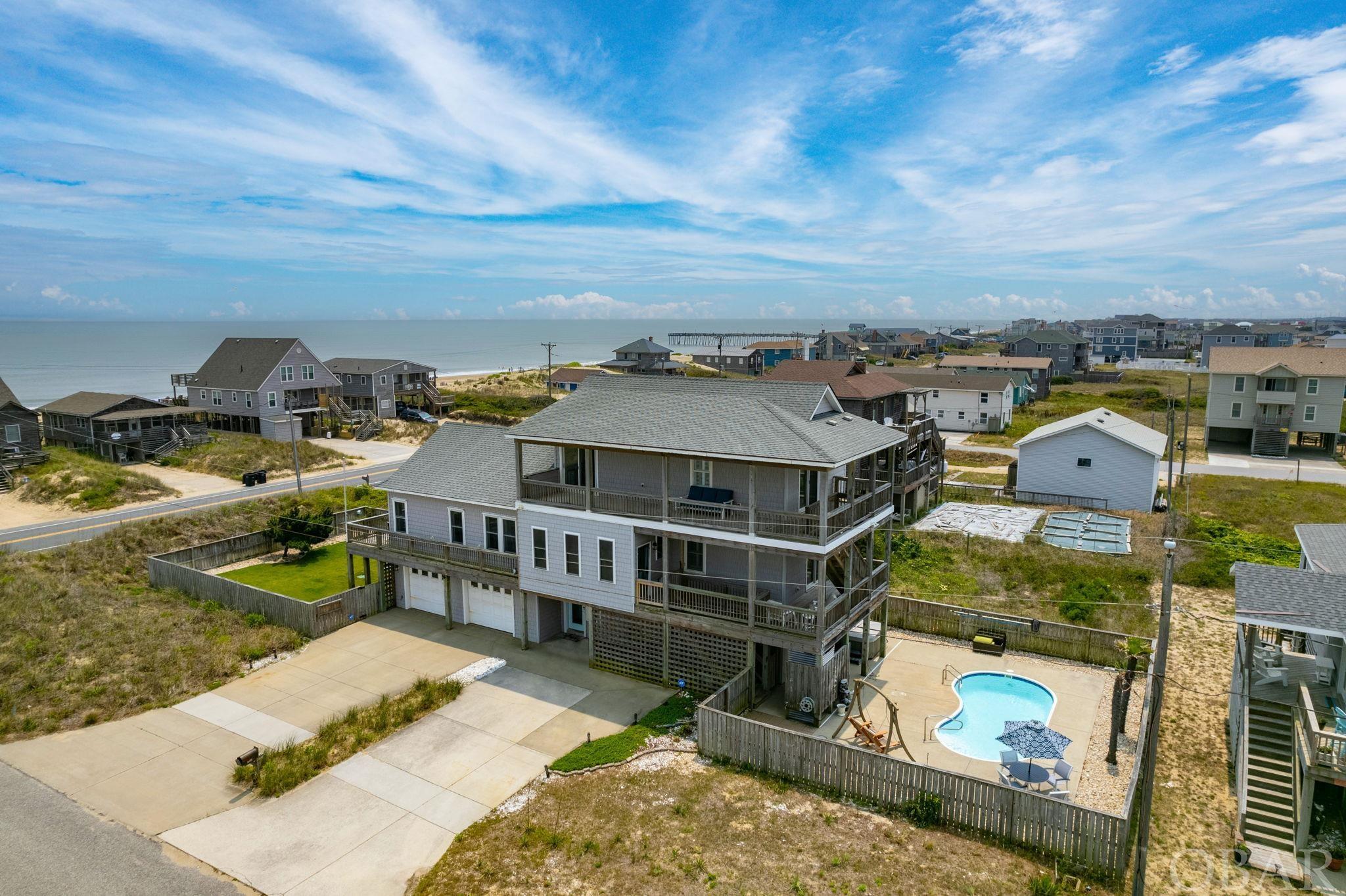 RARE opportunity semi-ocean front in the heart of Kill Devil Hills! From the moment you step on to the property, you will be captivated by the expansive ocean views, thoughtful design, and unique qualities. Situated on a corner lot, this home features an oversized landscaped and irrigated yard, a separate pool area complete with a brand-new hot tub. A new pool heater was installed in 2022, allowing you a longer swimming season. Two new HVAC units were also installed in 2022. The home is meticulously maintained and has something for everyone to enjoy. This would be a wonderful year around, second home, or investment opportunity. There is no shortage of storage space with the 2-car garage having plenty of room for all your beach toys. Upon entering you are greeted in a tiled entryway giving way to the ground level bonus room. This room is set up with 2 twin over double bunk beds, has its own fireplace, wet bar, and private ensuite. Up the stairs you'll walk into a bright and airy open living concept with custom plantation shutters and breathtaking ocean views. Vaulted ceilings and expansive windows allow so much natural light to pour in. The open living area is perfect for entertaining or just relaxing on the couch by the fire. Hardwood floors span this entire level going into the large primary bedroom equipped with an ensuite with a custom tile shower. Just off the kitchen, there was a screened porch that was recently converted to an all-seasons room in early 2023. This is an excellent addition for another seating area or office space. The third level houses the other 2 bedrooms and a jack and jill bathroom. All new furniture was purchased in 2021 and it is beautifully decorated. This home has wrap-around porches to sit and enjoy the sunrise over the ocean or just take in that fresh ocean air. The yard is fully enclosed and fenced, perfect for your fur babies. Other features include an outdoor shower, foot rinse, and on-demand tankless water heater. The septic was pumped and risers installed in 2021. There is so much to love about this house, you can tell every detail was well thought out. Don't miss your chance to tour it today!